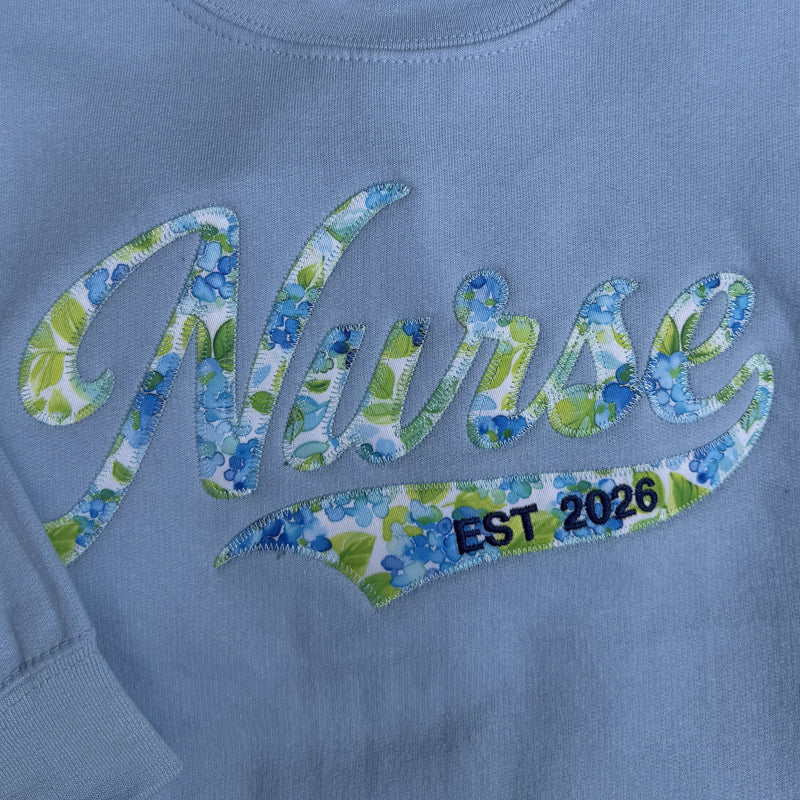 Blue sweatshirt with floral 'Nurse' text and 'EST 2026' on a matching blue background